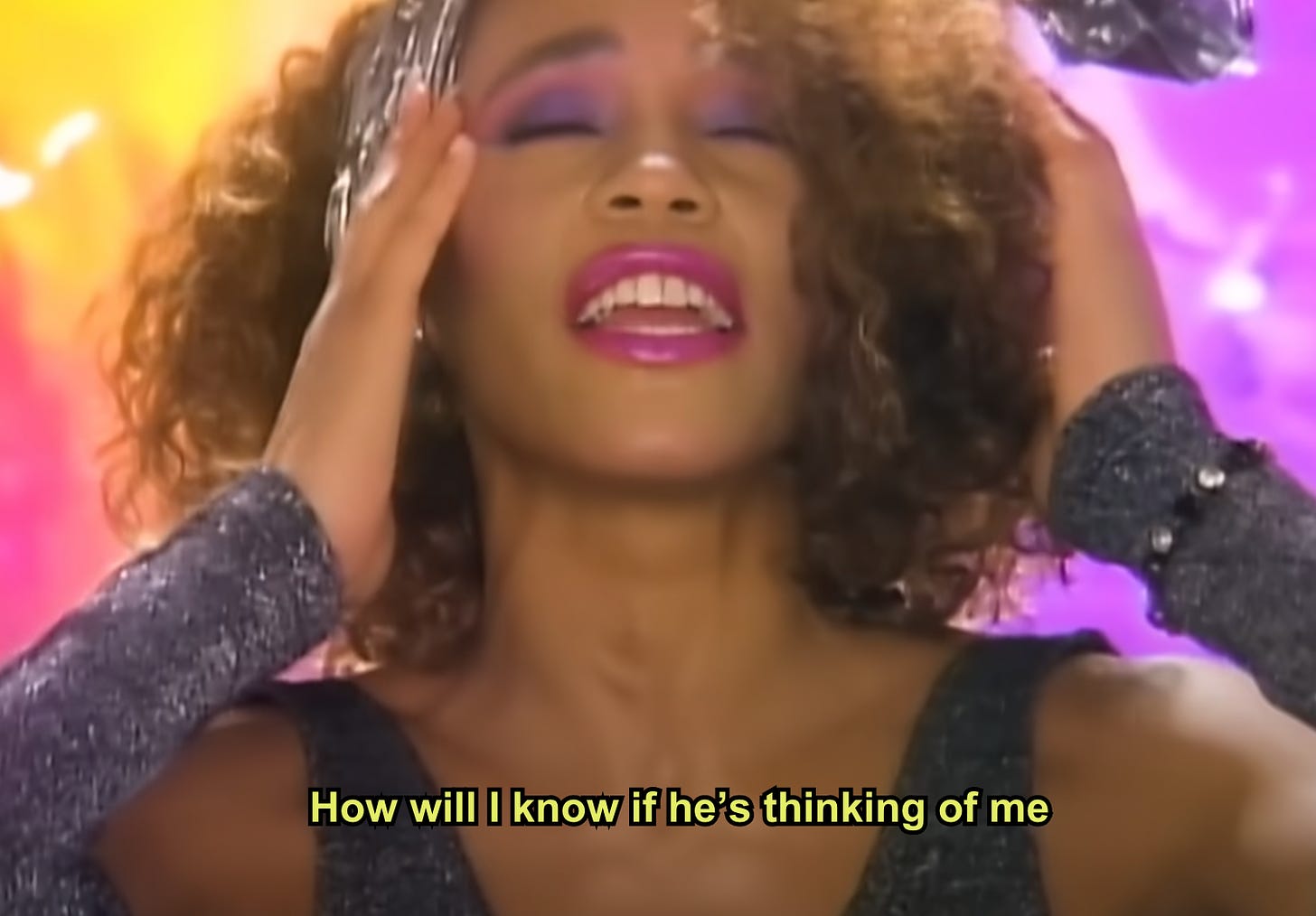 a still from the Whitney Houston music video "How Will I Know" showing her holding her head with her hands and eyes closed towards the sky with the lyrics "How will I know if he's thinking of me" in yellow text on the bottom