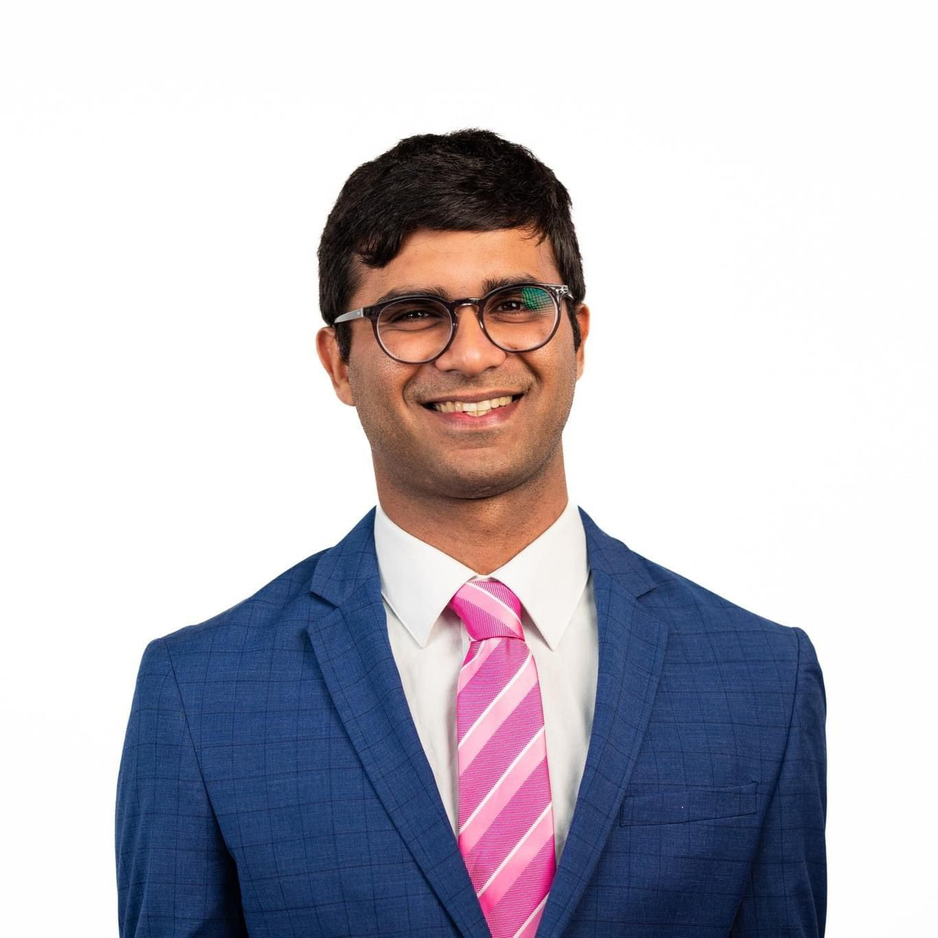 Aman Misra, a young Indian man with short black hair, is wearing glasses and smiling into the camera. He is dressed in a blue checked blazer, white shirt and pink striped tie.