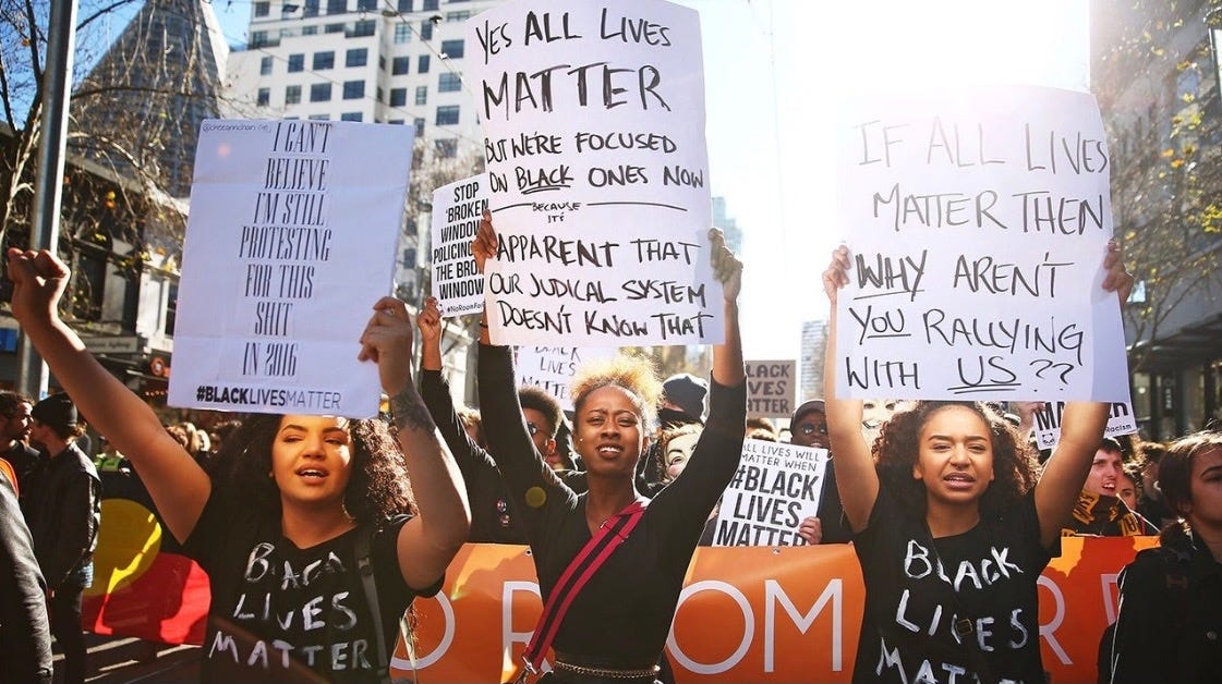 Three young Black women march in a protest. They each hold a sign above their head. Their signs read:   “I can’t believe I’m still protesting this shit in 2016 #BlackLivesMatters”  “Yes all lives matter but we’re focusing on the Black ones now because it’s apparent that our judicial system doesn’t know that.”  “If all lives matter then why aren’t you rallying with us?”