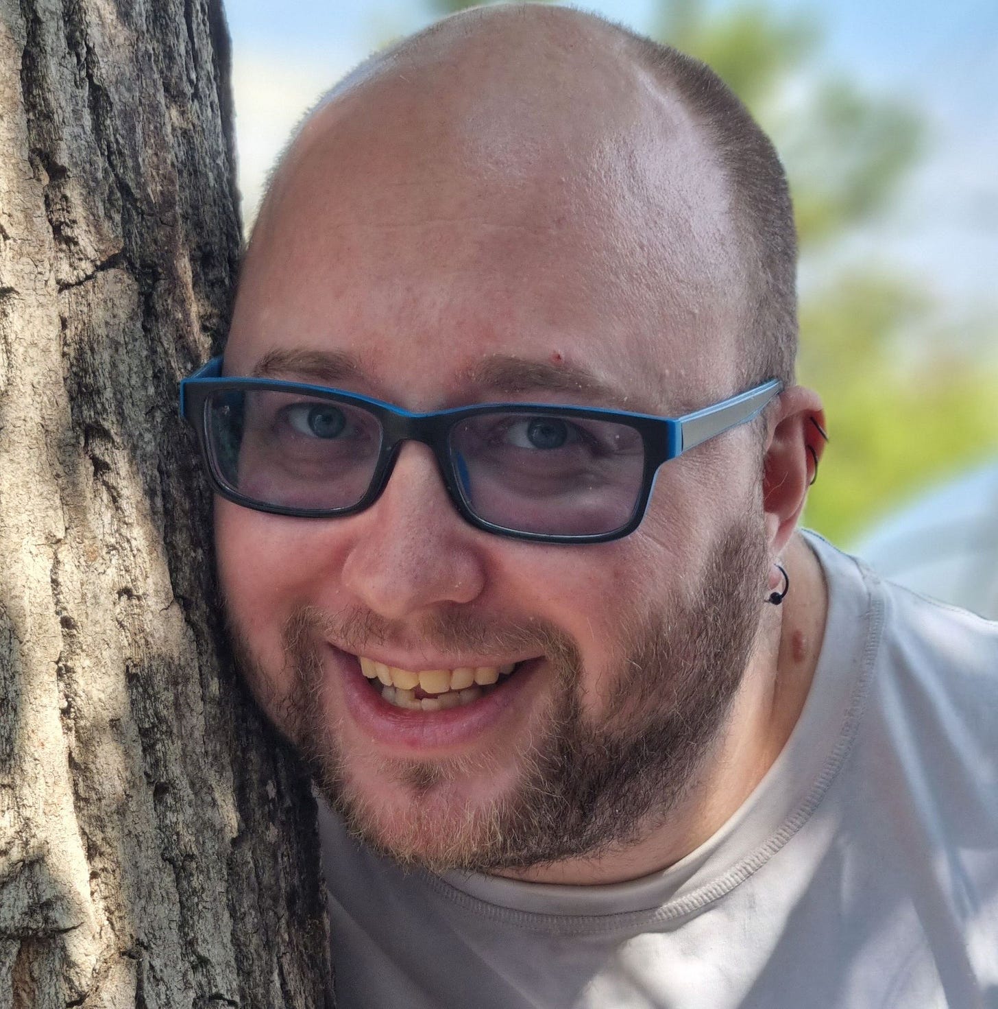 An agender person wearing glasses, with a bald top of the head and a light brown beard, just grown past the stubble stage.