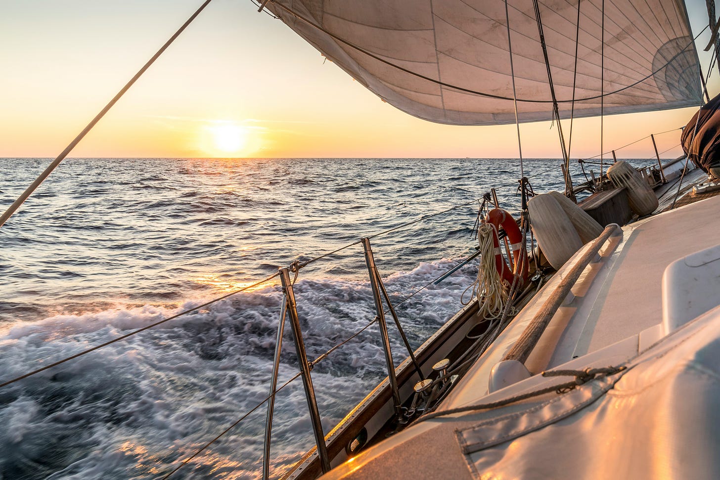 Image of the deck of a sailboat with the ocean and sunrise just off the port bow. 