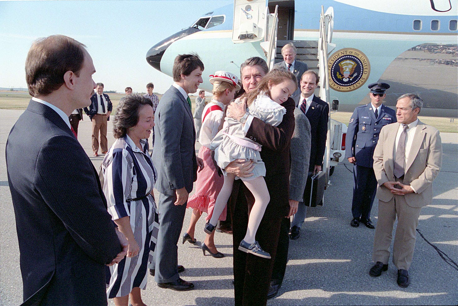 A sunny day on a tarmac in front of a light blue jet plane. Ronald Regan hugs a young Katie Beckett while looking at her father. Julie Beckett stands to the President's left in a navy and white jumpsuit. Several other people stand around in the background.