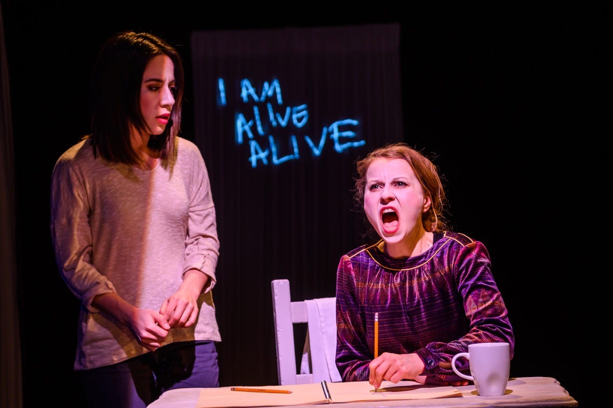 The words I AM Alive ALIVE in blue on black background behind two women, one standing on the left looking at the one seated at a small flat table/desk, who is screaming the words "I am alive" her left hand holding a pencil as she writes the words of a spirit speaking to her through the pencil.