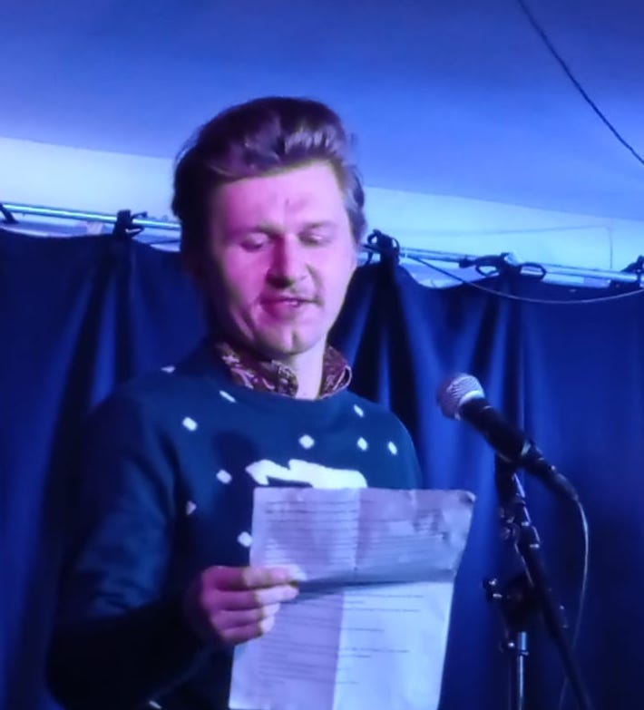 Christian stands on a stage, reading from a piece of paper. He is wearing a Christmas jumper and is lit by soft blue light. There is a mic in front of him and stage curtains behind him.