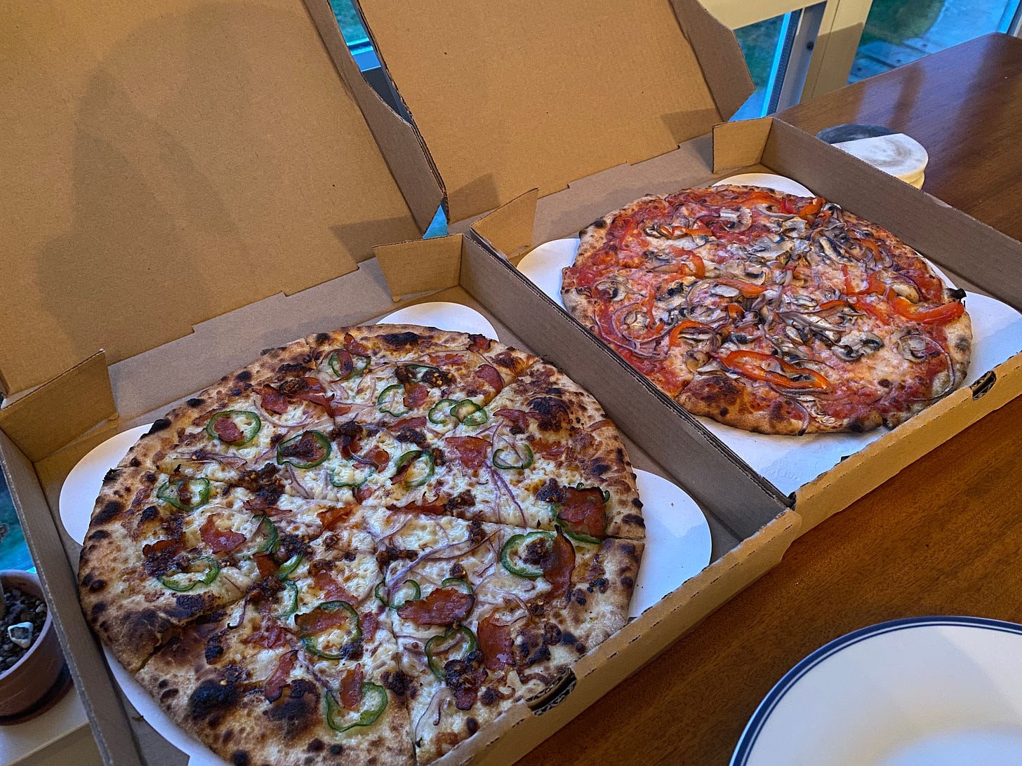 Two open cardboard pizza boxes beside each other, each with a thin-crust pizza inside. One has a sauce-free base and is covered with cheese, jalapeño, red onion, and torn slices of pepperoni. The other is a classic red sauce with mushrooms, red onion, cheese, and red peppers.