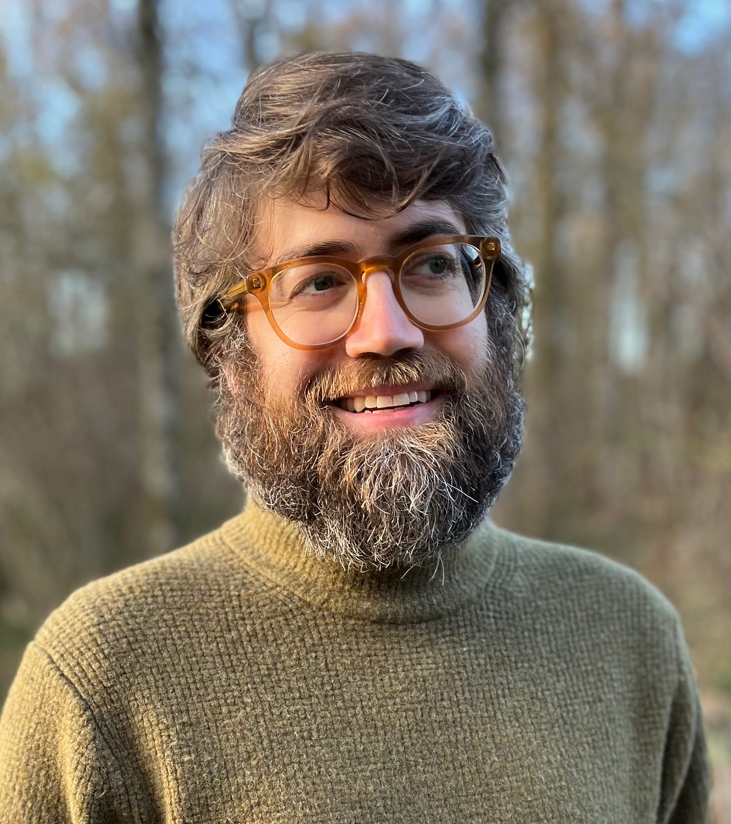 Photo of me, Zachary Dillon. I'm 39 years old, white, and I have glasses, a beard, and I'm wearing a green sweater. I'm standing in the woods, so there are blurry trees behind me. Oh, and I'm smiling, because I like the woods, not because I like having my picture taken.