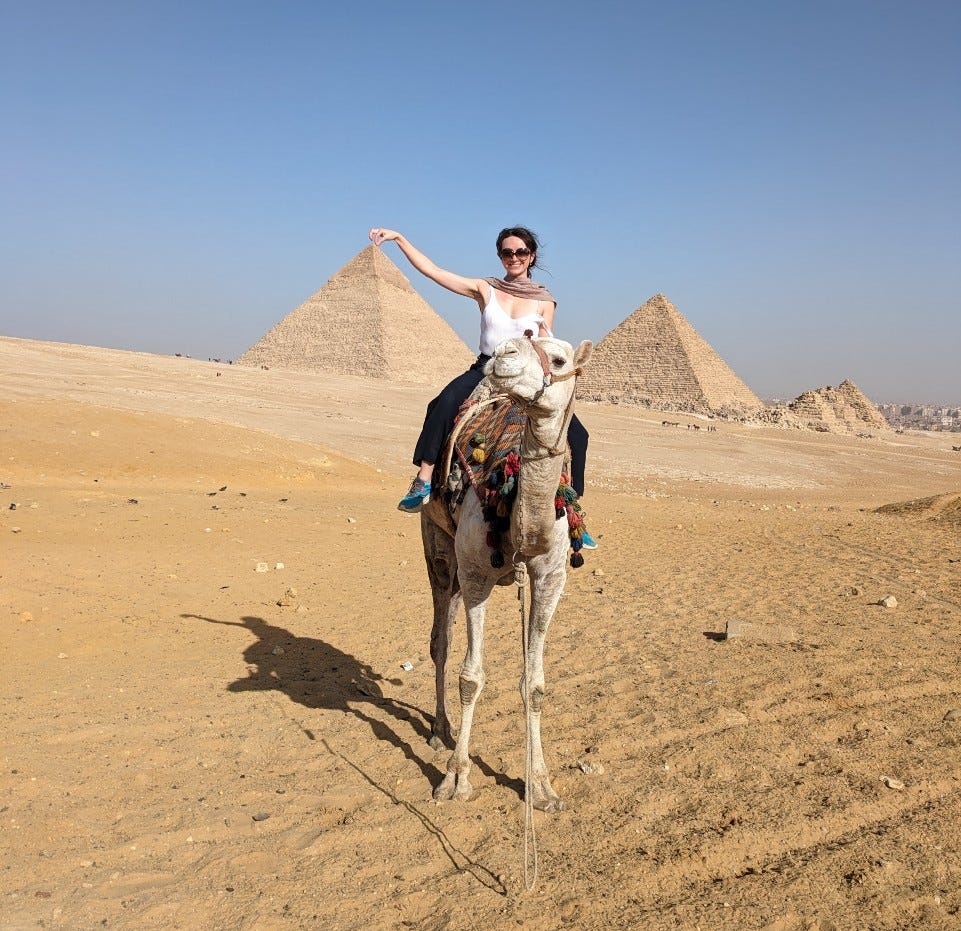 A white woman in sunglasses rides a camel, pyramids in background