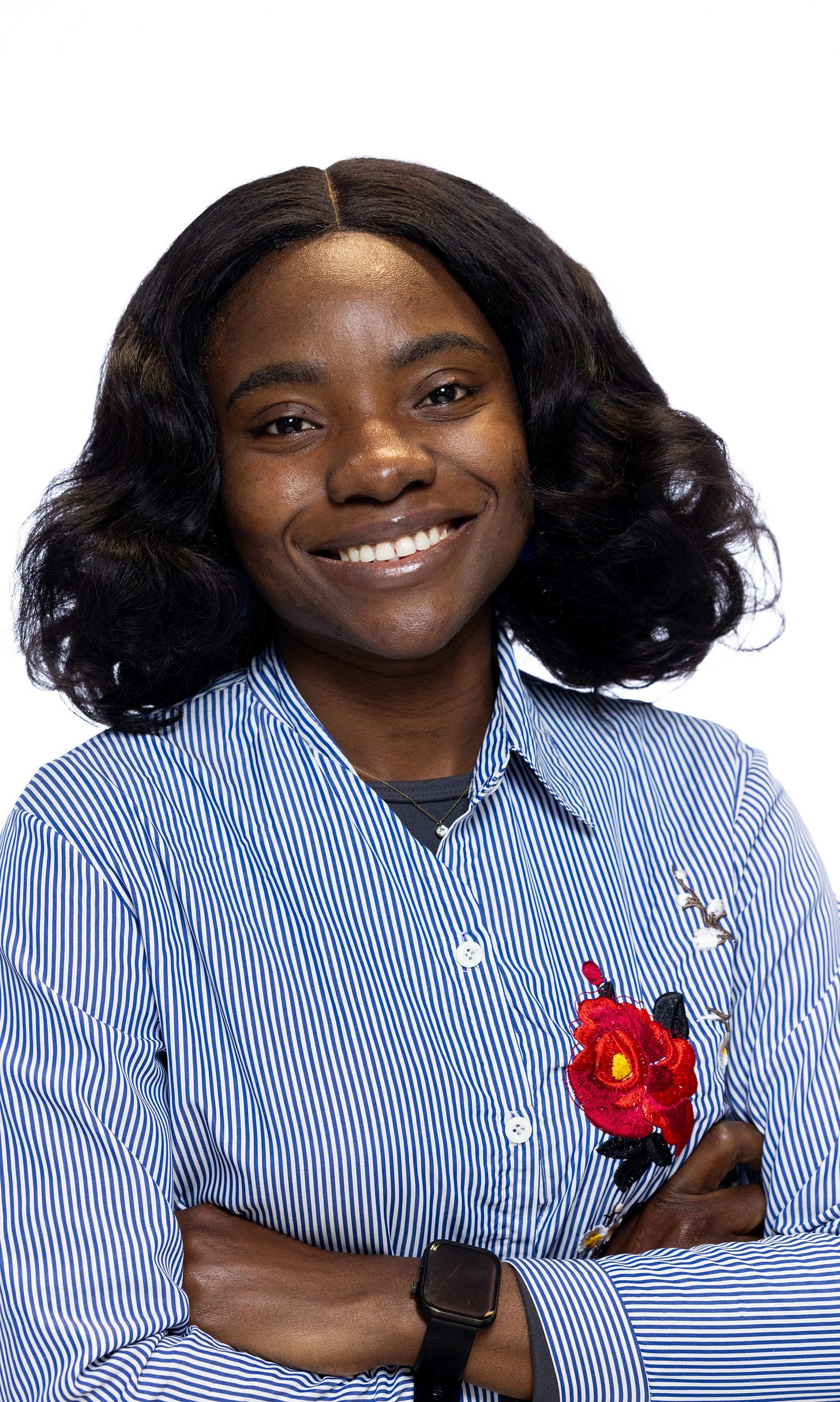 Zainab is a Nigerian woman with short, black wavy hair almost touching her shoulders. She is wearing a blue striped shirt with an embroidered red rose on the left side and has a bright smile on her face. 