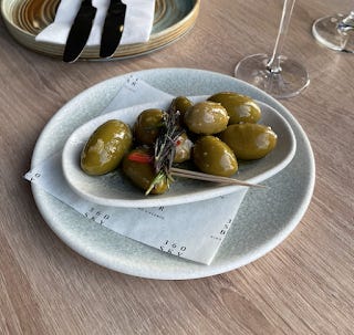 A white bowl on a white plate, filled with large olives, coated in oil, and flecked in slices of chilli.