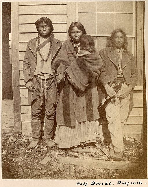 sepia photo of 3 Metis adults and a baby standing in front of a building.