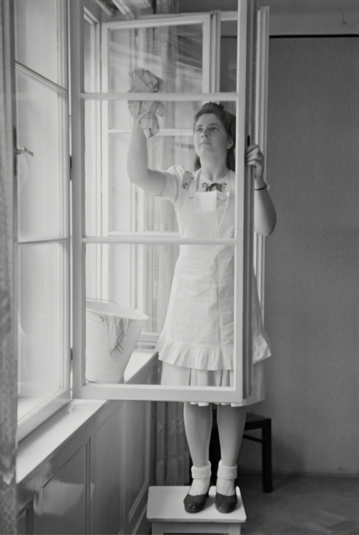 A black and white photo of from the 1940s of a person wearing a frilly uniform standing on a stool inside a room and cleaning a window with a rag
