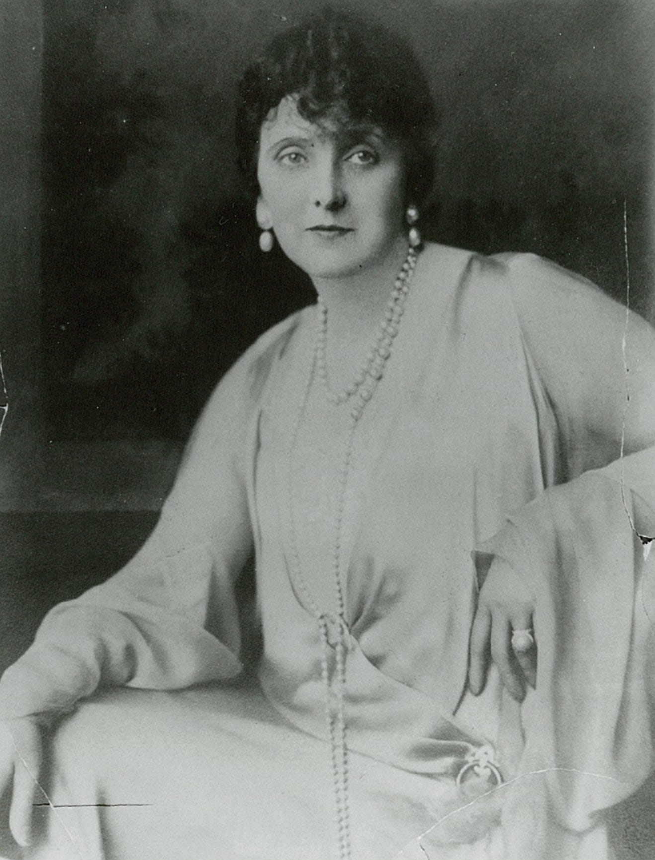 Emily Post, 1920, 47 years old. White woman, dark hair, heavily lidded eyes, approachably glamourous, large pearl drop earings, long strand of pearls doubled to two lengthes, white dress very 1920s style