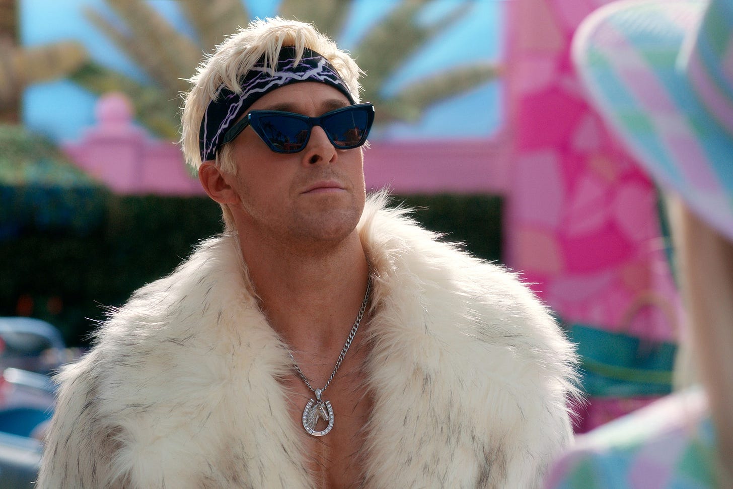 Ryan Gosling as Ken, wearing a faux fur coat, sunglasses, bandana, and a horseshoe necklace as he introduces Barbie to his Mojo Dojo Casa House.