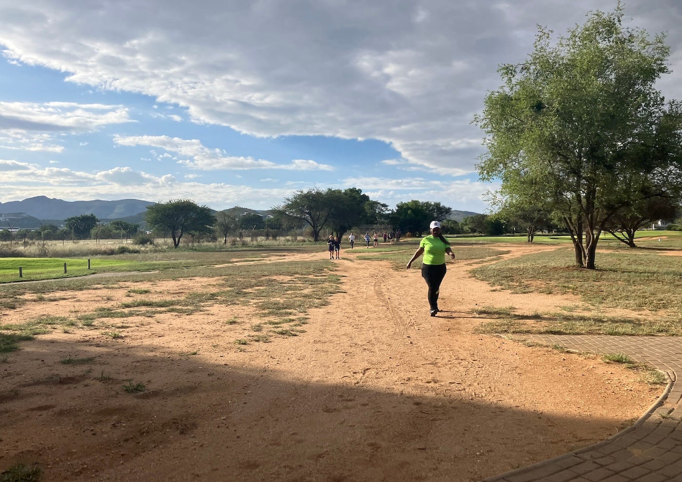 A wide sandy area with tufts of grass and a few people approaching, on the first half of their parkrun