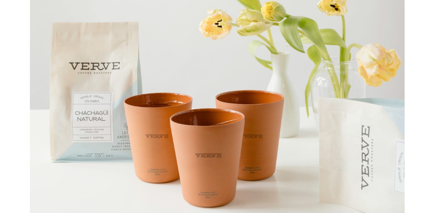 Three Verve-branded rust-colored clay cups sit on a white table next to a vase of yellow flowers and in front of a white Verve-branded coffee bag.
