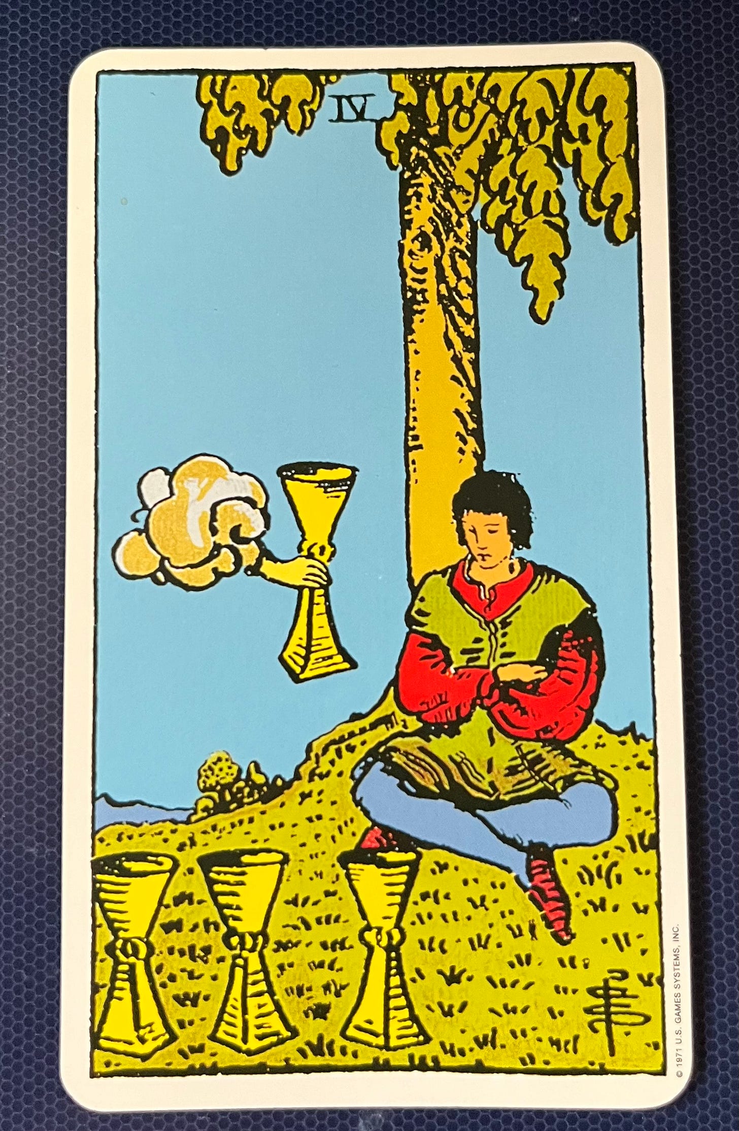 The image is of a tarot card, specifically the Four of Cups. It depicts a person sitting under a large tree with abundant foliage. The individual is holding one golden chalice and there are three additional chalices on the ground beside them. Above in the sky, there’s a cloud from which an arm extends offering yet another golden chalice to the individual. The background is blue with a white border around the edge of the card
