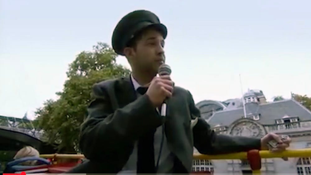 A screenshot from the apprentice, where one of the contestants is dressed as a tour guide and is sitting on the top deck of a bus talking to a crowd through a microphone