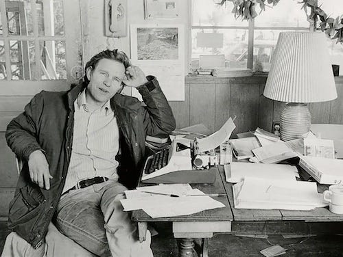 Photo of John Nichols, taken from NY Times obituary. Nichols is seated at his desk wearing a button down shirt, blue jeans and winter jacket (open). He looks at the camera with a friendly gaze. On the table before him there is a manual typewriter, a coffee cup, and several loosely stacked piles of paper. On the wall behind him are a telephone and some photographs.