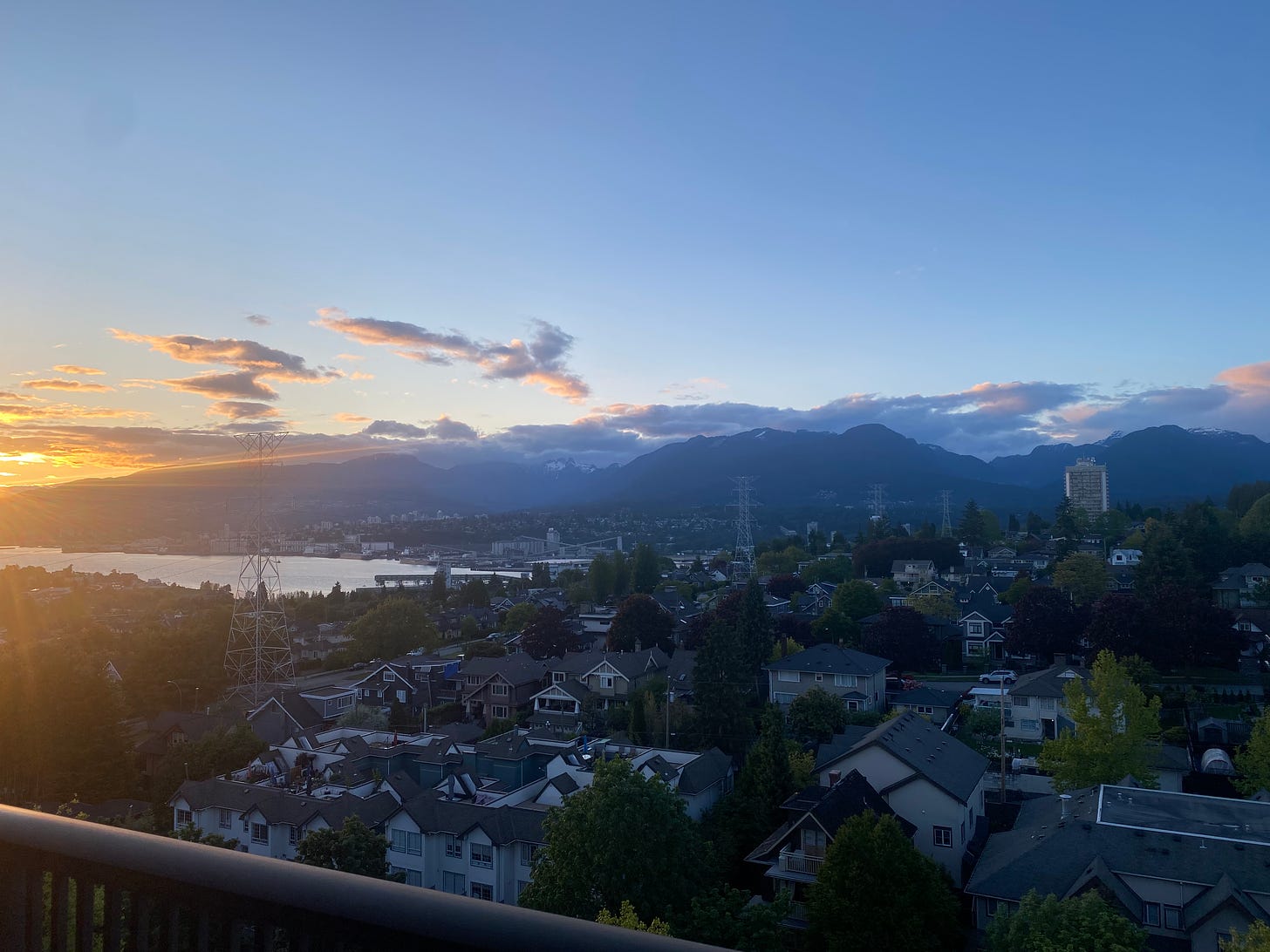 A view from north Burnaby at sunset, looking over the inlet and mountains. There is a splash of gold from the sun in the left side of the frame, otherwise everything is very blue, including the sky and the houses in the foreground.