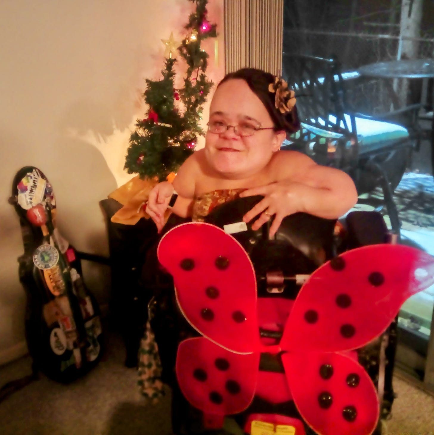 Gaelynn is seated backwards in her electric wheelchair to show off the red and black ladybug wings that are attached to the handlebars of her chair. She is smiling. She is wearing a yellow and orange dress and her brown hair is pulled up with a yellow flower clip. Behind her is a small Christmas tree and a garland with colored Christmas lights. Her violin case is propped against the wall.