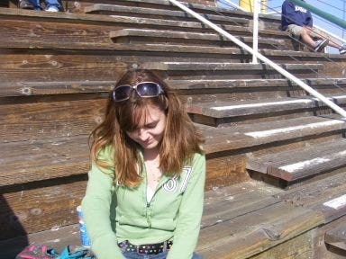 a teenage woman looks shyly down and away from the camera. she is seated on a set of wooden bleachers at the edge of the pacific ocean.
