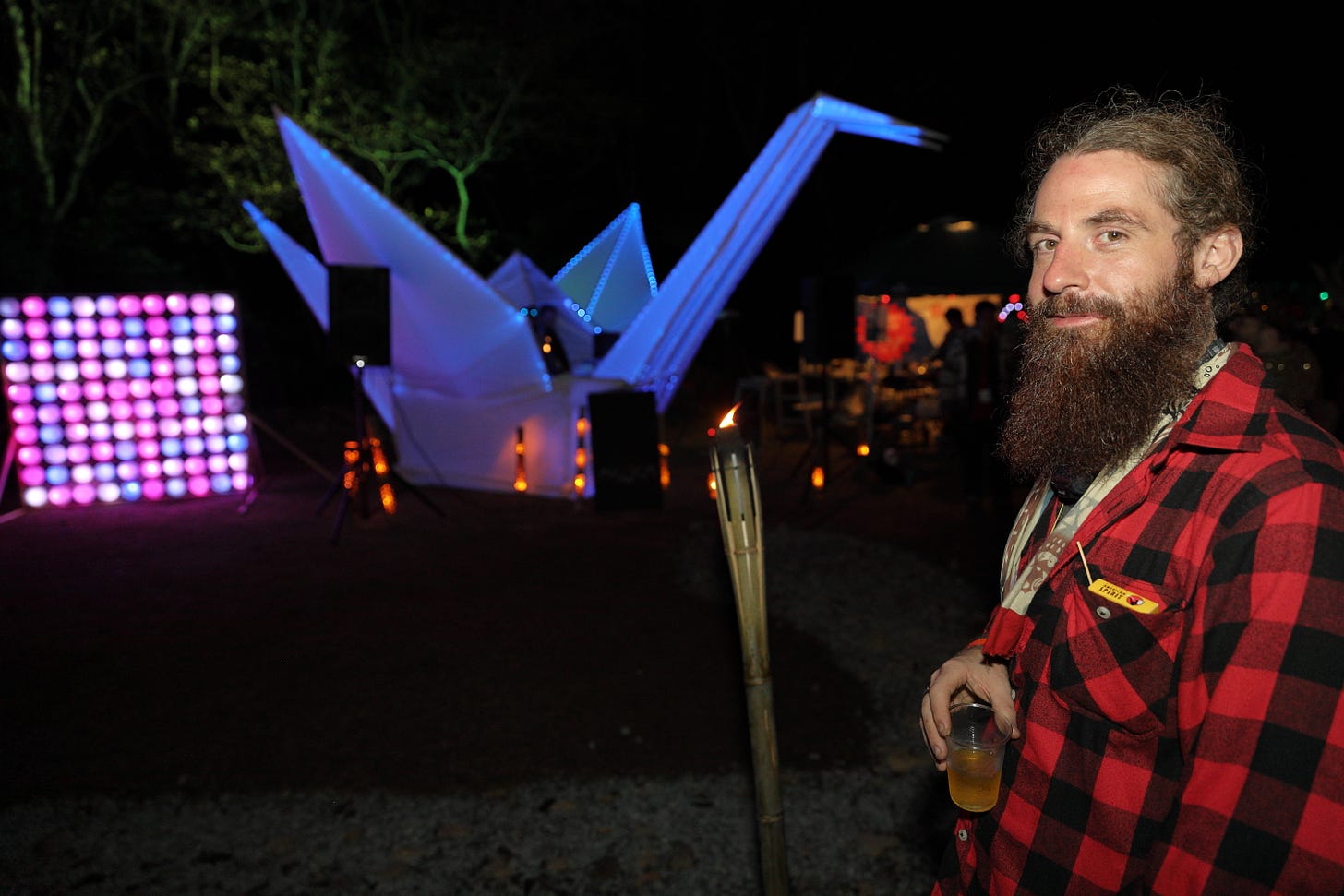 Matthew Ketchum in a red and black plaid long sleeve shirt with a drink smiling at the camera at Burning Japan 2023. Three headed neon crane in the background, night.