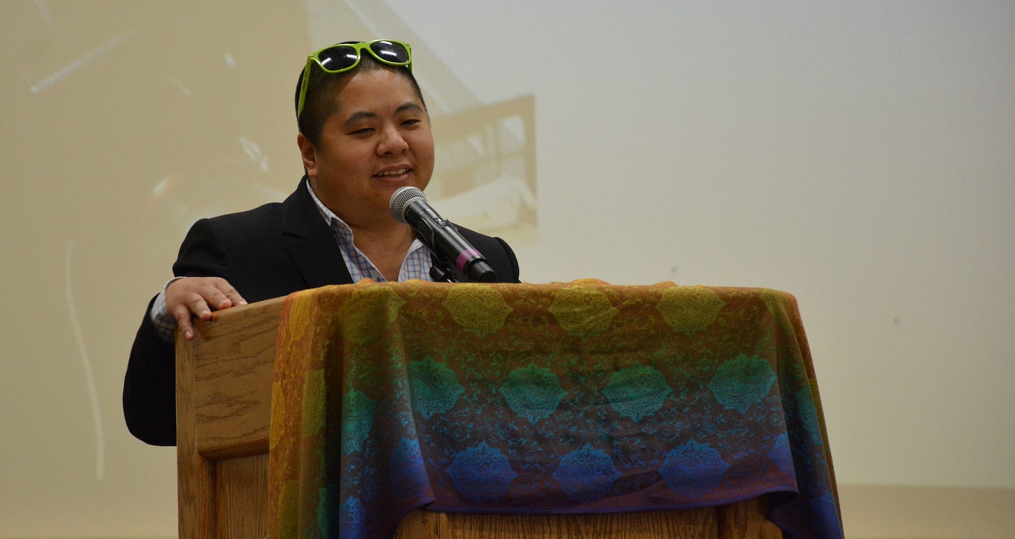 Ching-In Chen, smiling a little, speaks into a mic at a podium draped with multi-colored They are wearing a suit jacket and sunglasses on their head. 