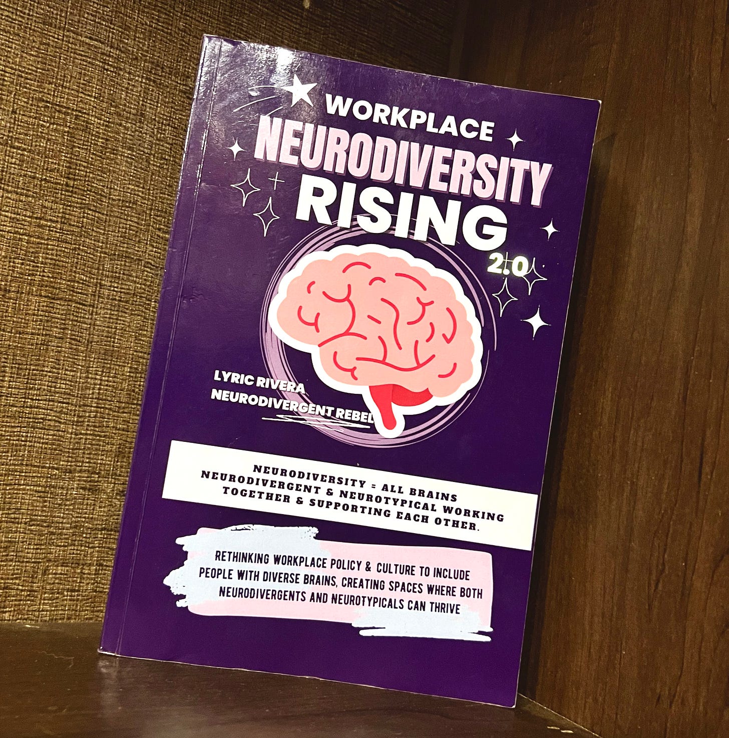 A purple book with a pink brain on the cover, titled Workplace NeuroDiversity Rising 2.0, by Lyric Rivera, sits on a shelf in the corner.