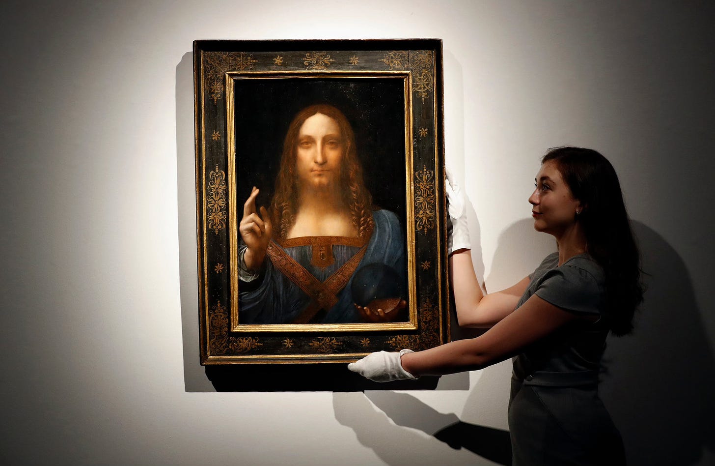 A young woman with white gloves holds Leonardo Da Vinci's “Salvator Mundi” (1500), a painting of Christ set in a black and gold frame.