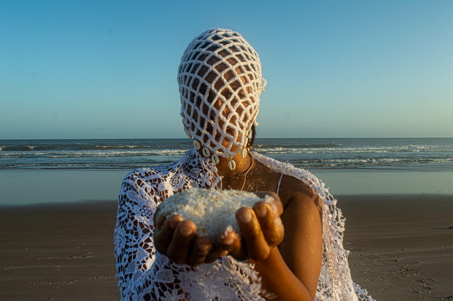 person wearing a white mask decorated with cowry shells holding salt by the ocean