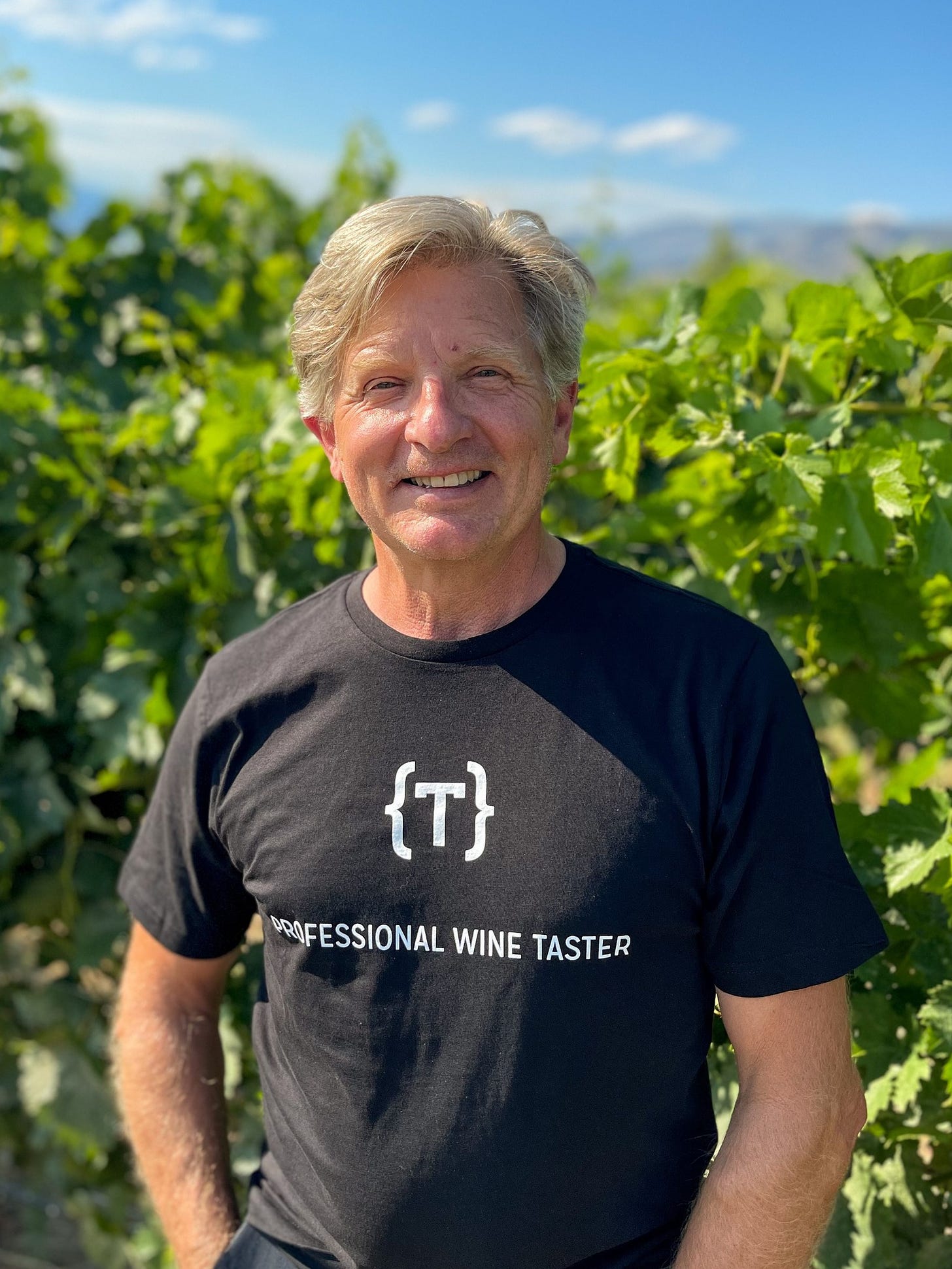 A winemaker stands for a portrait shot, the soft focus background is a green vineyard with a blue sky.