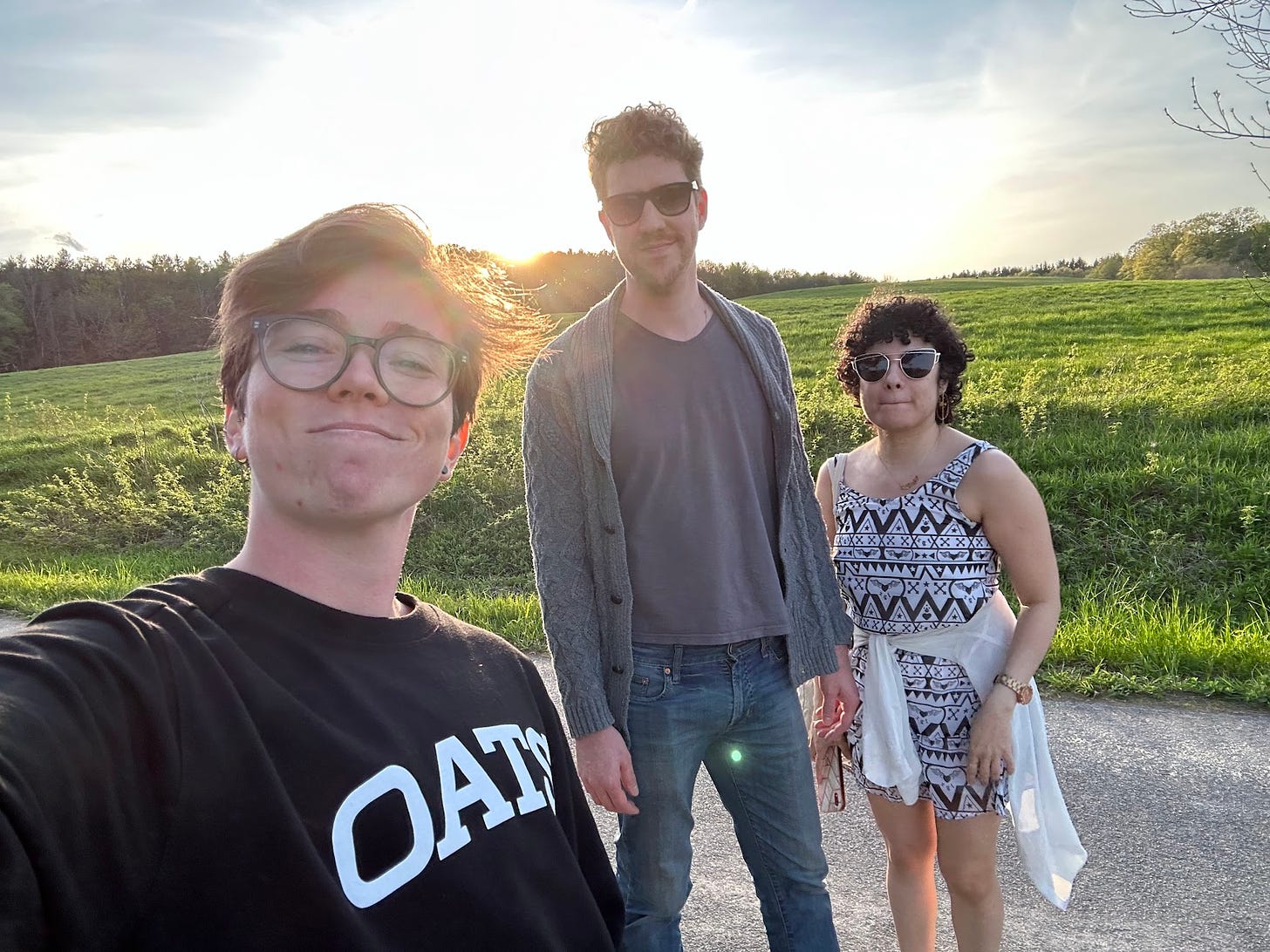 Three people smile at the camera standing on a paved road in front of a field of grass where the sun is setting