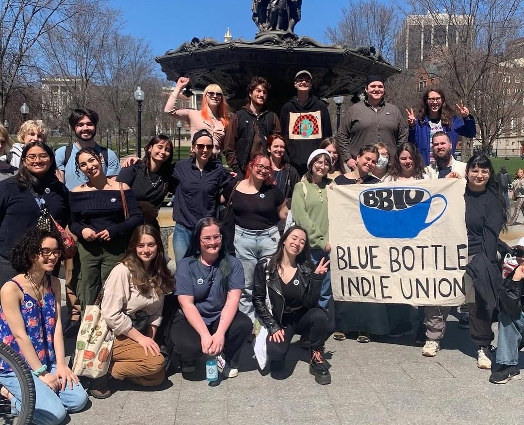 A group of BBIU workers pose in front of fountain and display poster reading "BBIU Blue Bottle Indie Union."