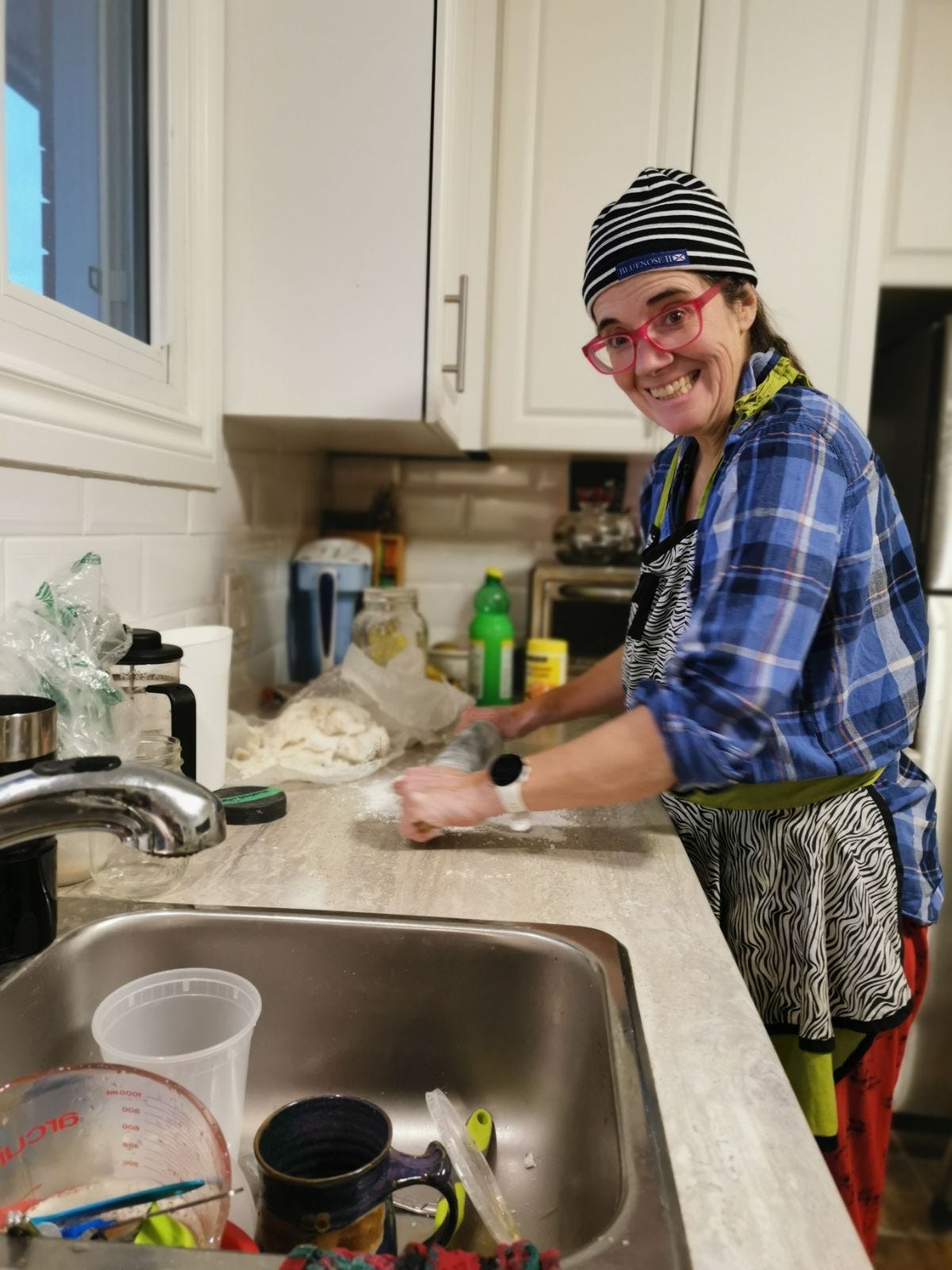 A white woman with a silly smile, in a striped beanie, blue checked shirt, zebra striped apron and red pyjama pants and pink eye glasses. 