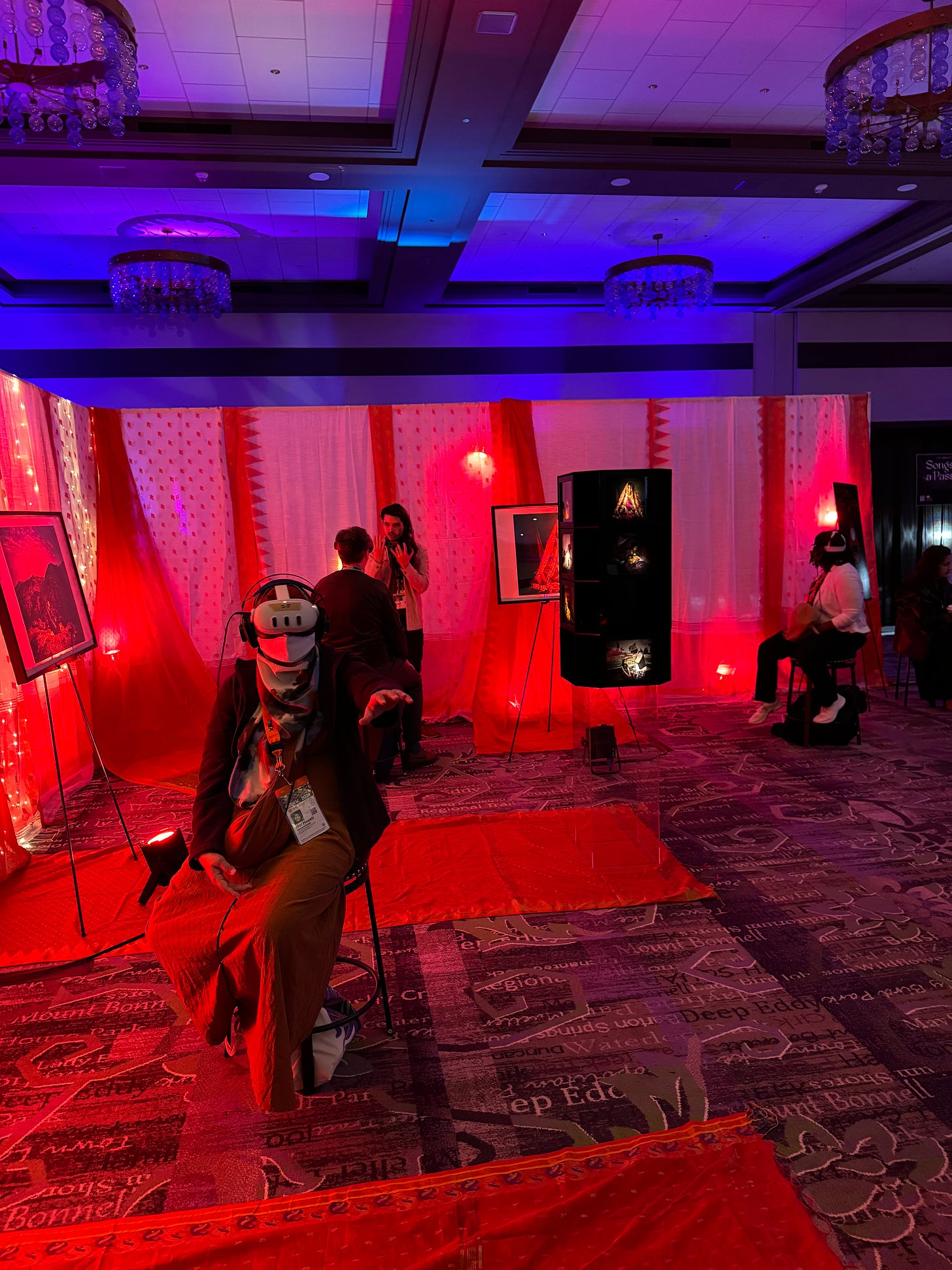 A few people sit on stools in a dimly lit exhibition hall for the XR exhibition. All wear XR headsets on their faces, and an eerie red/blue glow is in the background.