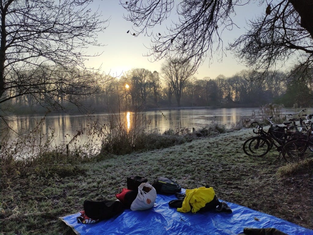 A low watery sun pokes through trees over a lake with a smattering of ice, with bikes and bags on the grass.