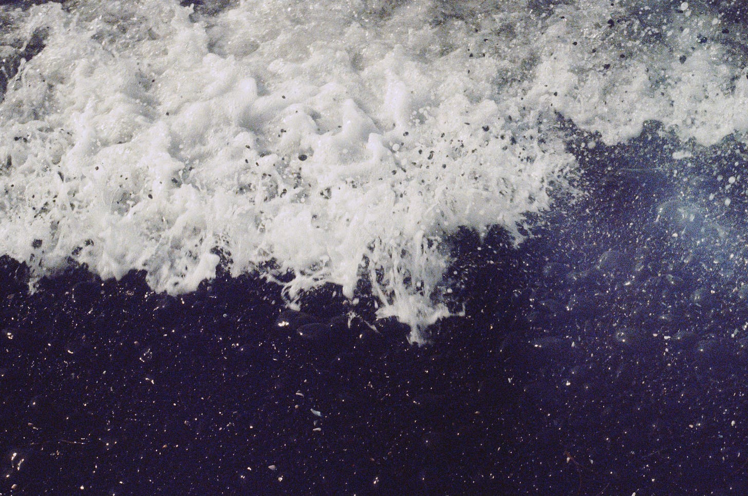 A close-up photograph of ocean water, white and frothy, flowing onto a rocky shore.