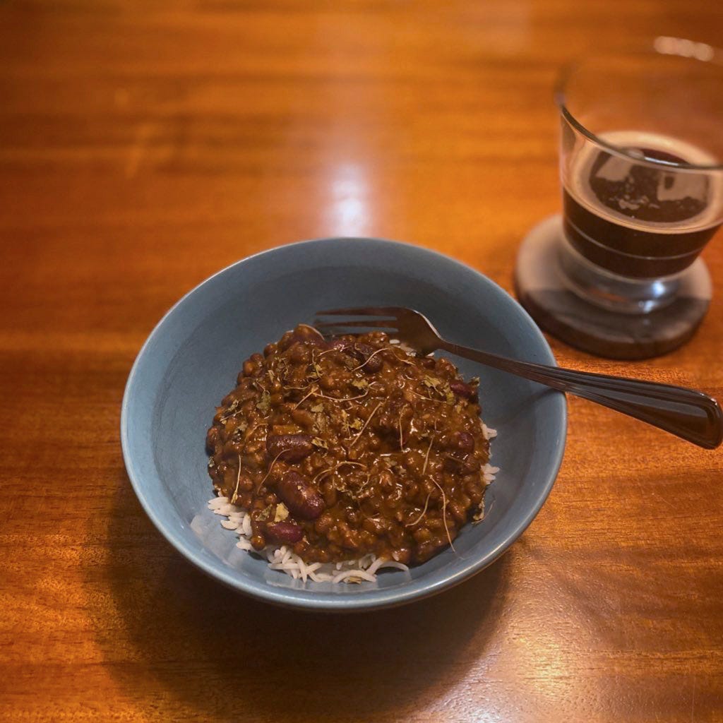 A blue bowl of white basmati rice with deep brown dal on top, sprinkled with dried fenugreek leaves. A fork rests at the back of the bowl and there is a partially-full glass of dark beer on a coaster above and to the right.