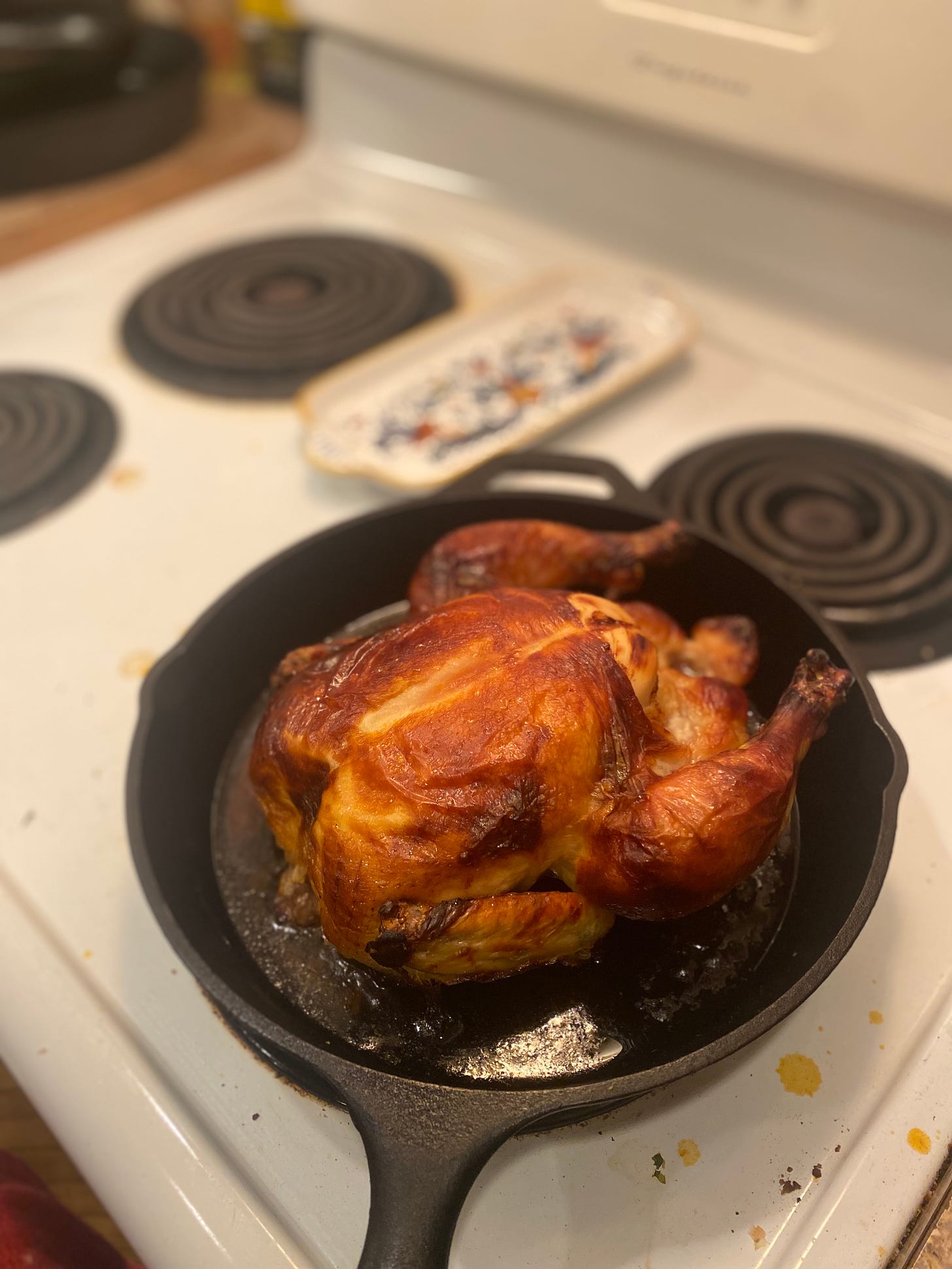 In a cast iron pan is a roast chicken with crispy, caramelized skin, the drippings shining in the bottom of the pan. The stove it's resting on is a bit messy.