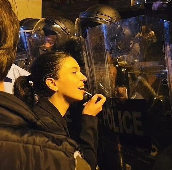 A Georgian girl stands in front of a line of riot police, using the reflection in their riot shield to apply her makeup.