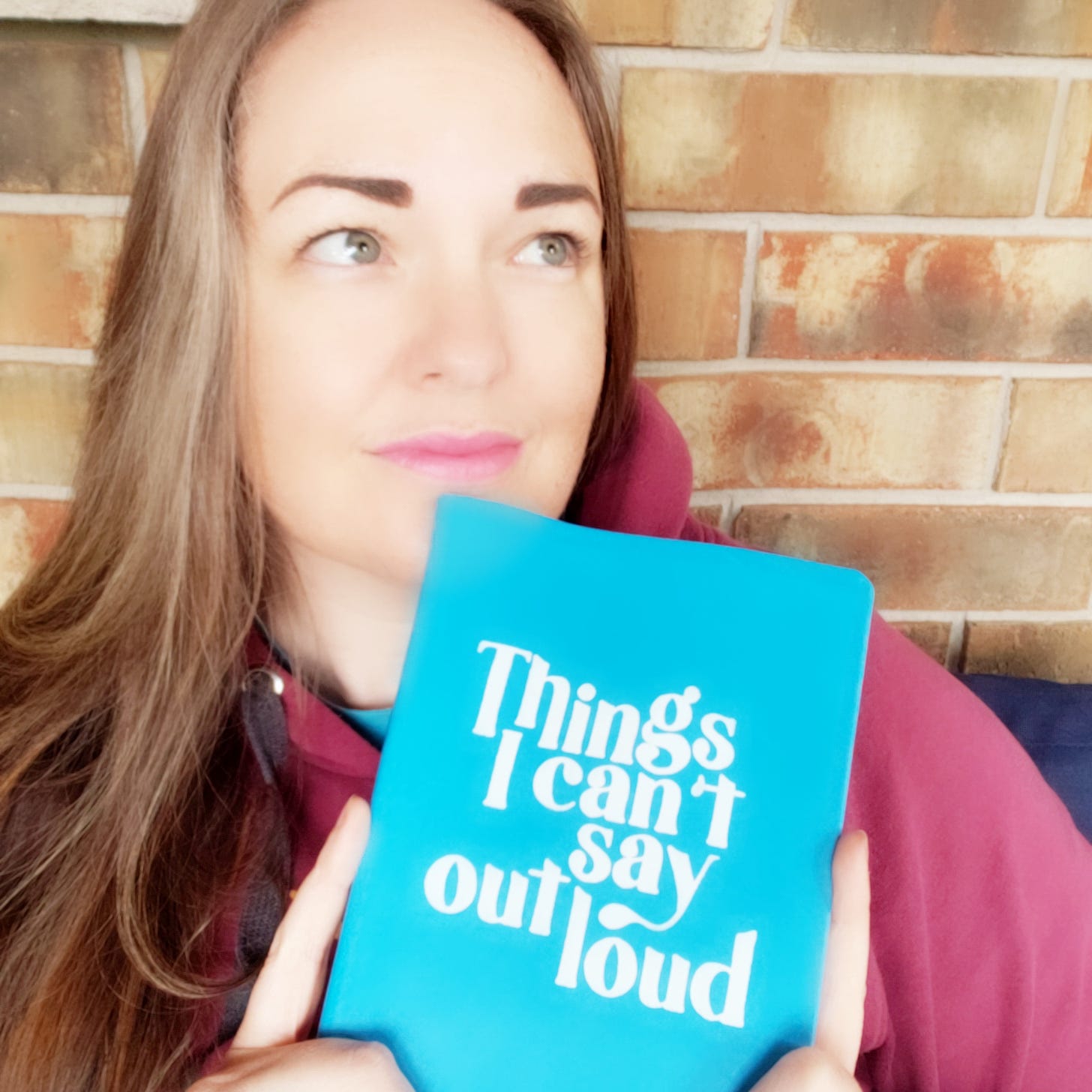 a white woman, me, sits in front of a brick wall and holds a book with the cover "Things I can't Say Out Loud"