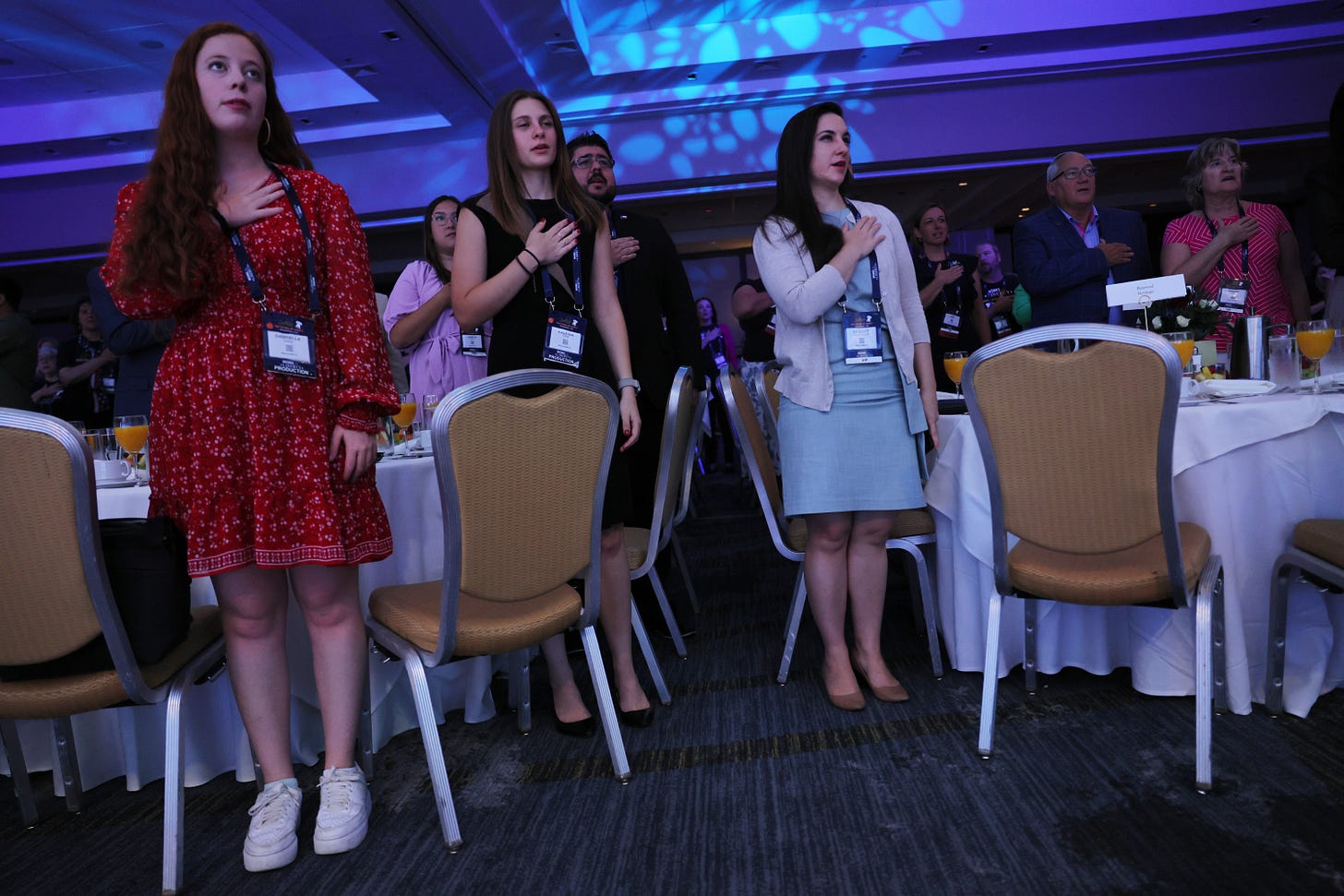 Three young white women stand in a conference room, facing the same way with their hand on their heart, at a recent "Moms for Liberty" conference