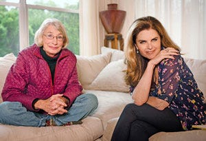 Mary Oliver sits cross-legges on a couch. She is wearing thin-framed glasses and a pink puffy coat, jeans, and socks. Next to her sits Maria Shriver, posing elegantly in a floral blouse and dark slacks.