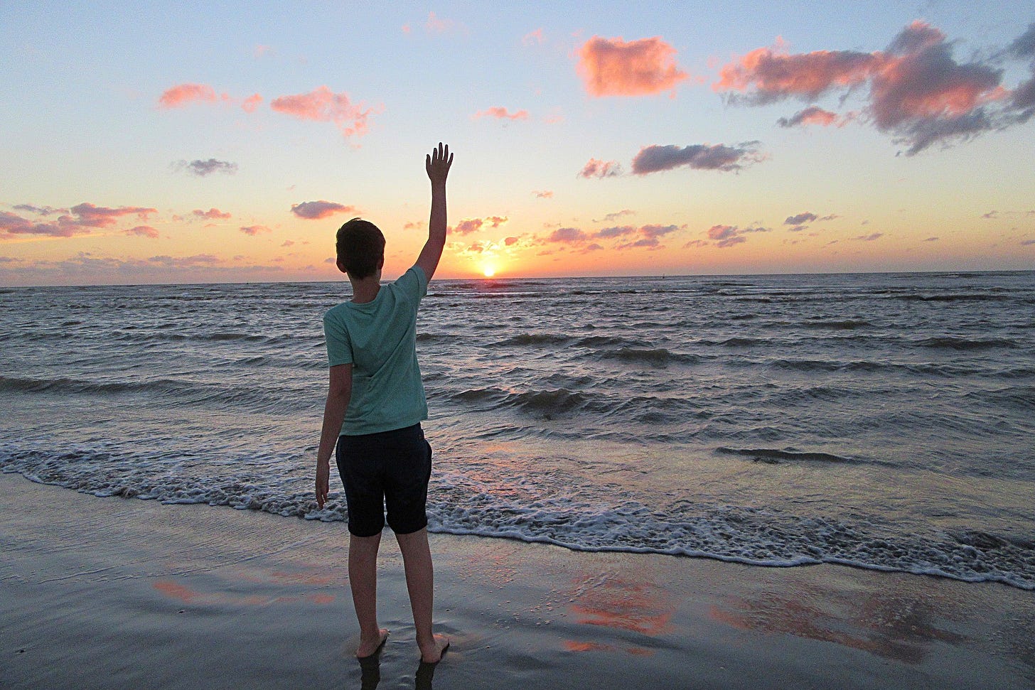 A young boy stands on the shore waving goodbye to a setting sun.