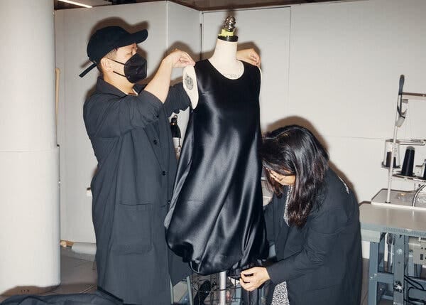 Mr. Do, in his long black jacket, ball cap and mask, adjusts the armholes of a black dress on a mannequin. A woman in a black jacket pins the hem. 