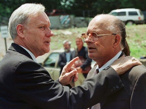 Mr. Pepper, wearing a dark suit, stands in the open air with his right hand on the shoulder of a similarly dressed man with glasses and thinning hair.