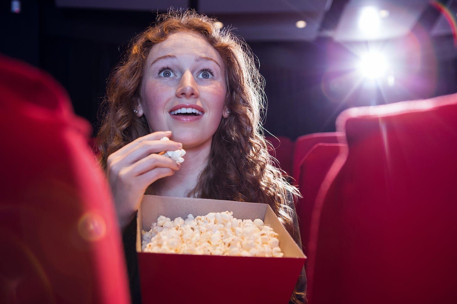 Young woman with long red hair, eating popcorn, looking amazed in a cinema filled with red seats