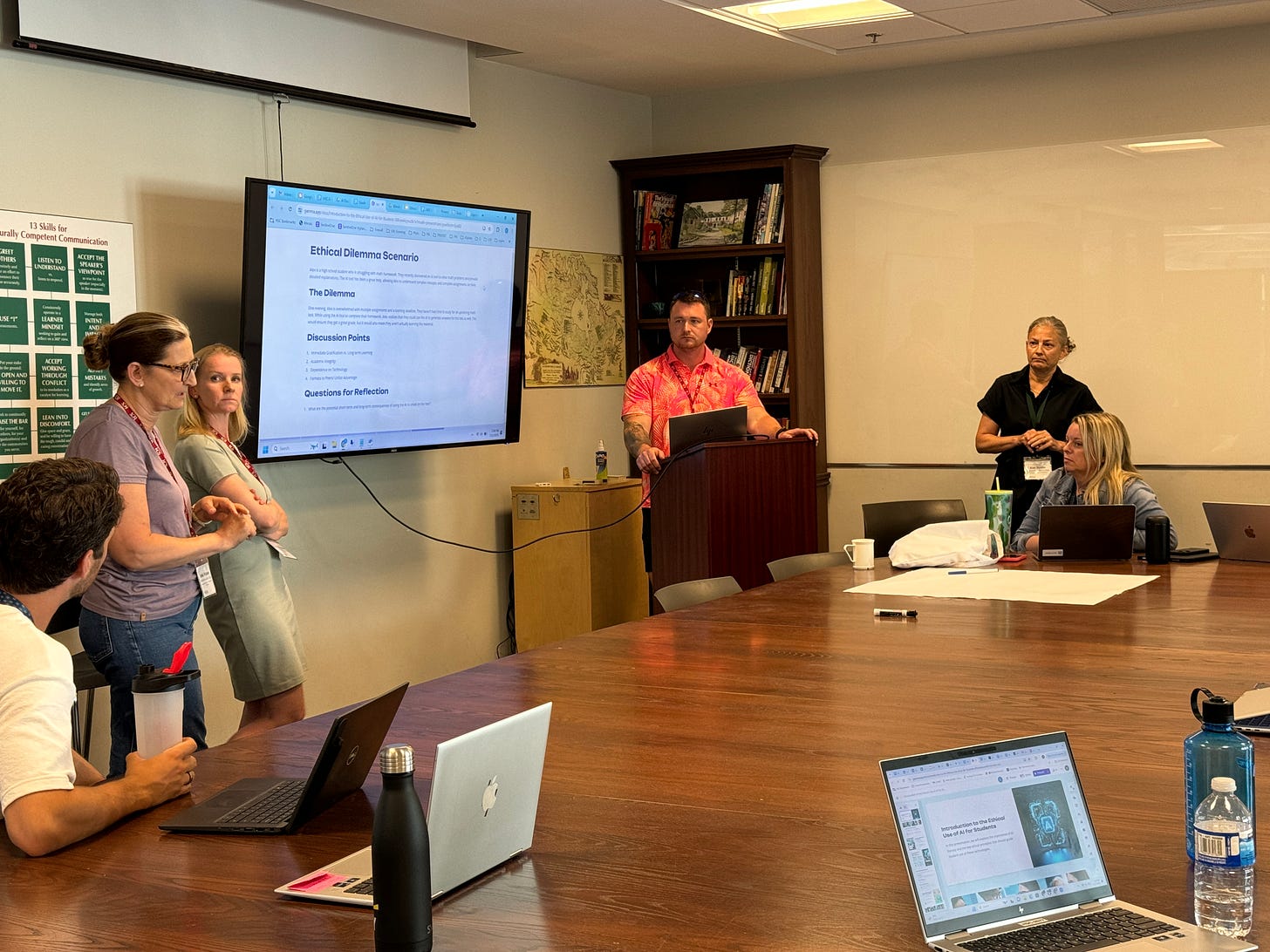 A group of people is participating in a meeting in a conference room. One man stands at a podium, presenting information displayed on a large screen titled "Ethical Dilemma Scenario." Two women stand to the left of the screen, one speaking and the other listening. Several others are seated at the table with laptops and water bottles. 