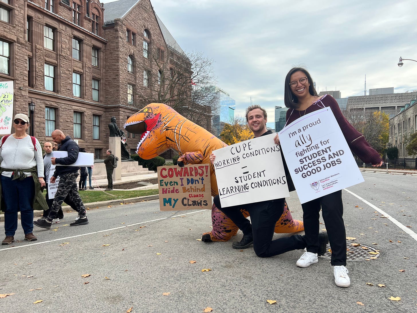 My partner and I standing next to the Provincial Parliament with signs in support of education workers. There is also a person in a dinosaur costume next to us.