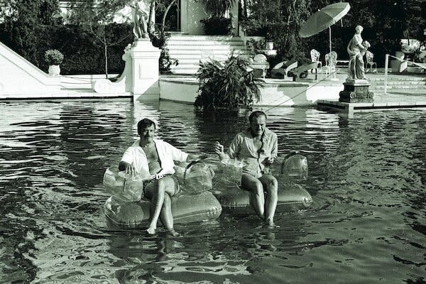 A black-and-white photo of the actors Eli Wallach and David Niven sitting on inflatable chairs on the water.